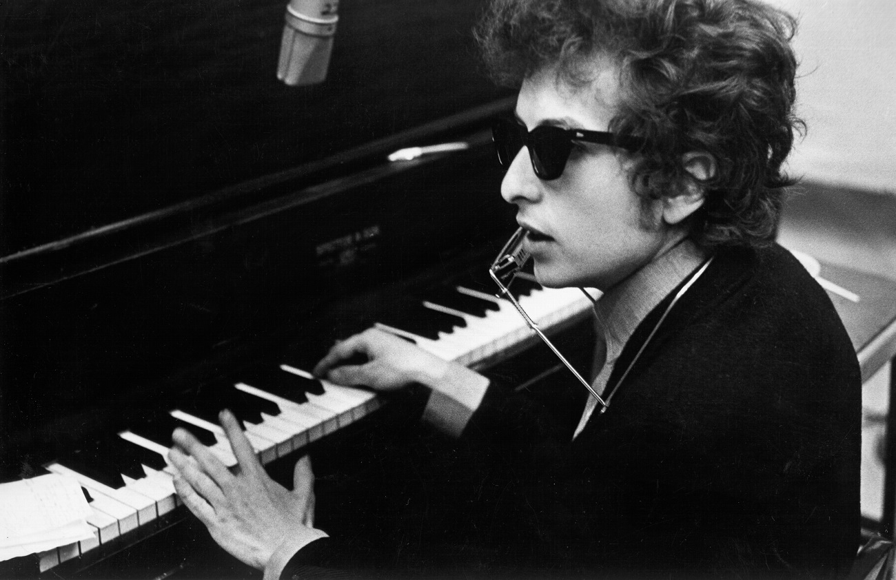 A black and white picture of Bob Dylan wearing sunglasses and sitting at a piano.