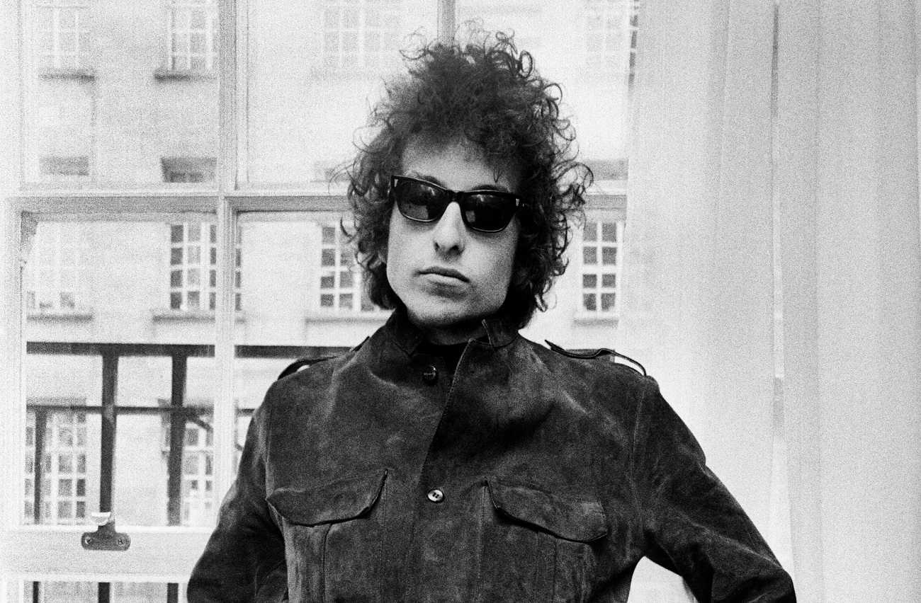 A black and white picture of Bob Dylan wearing sunglasses and sitting in front of a window.