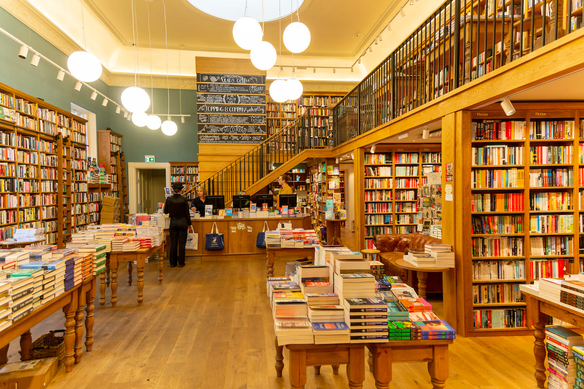 An interior shot of a bookstore with bookshelves, filled to the brim with books and a customer making a purchase.