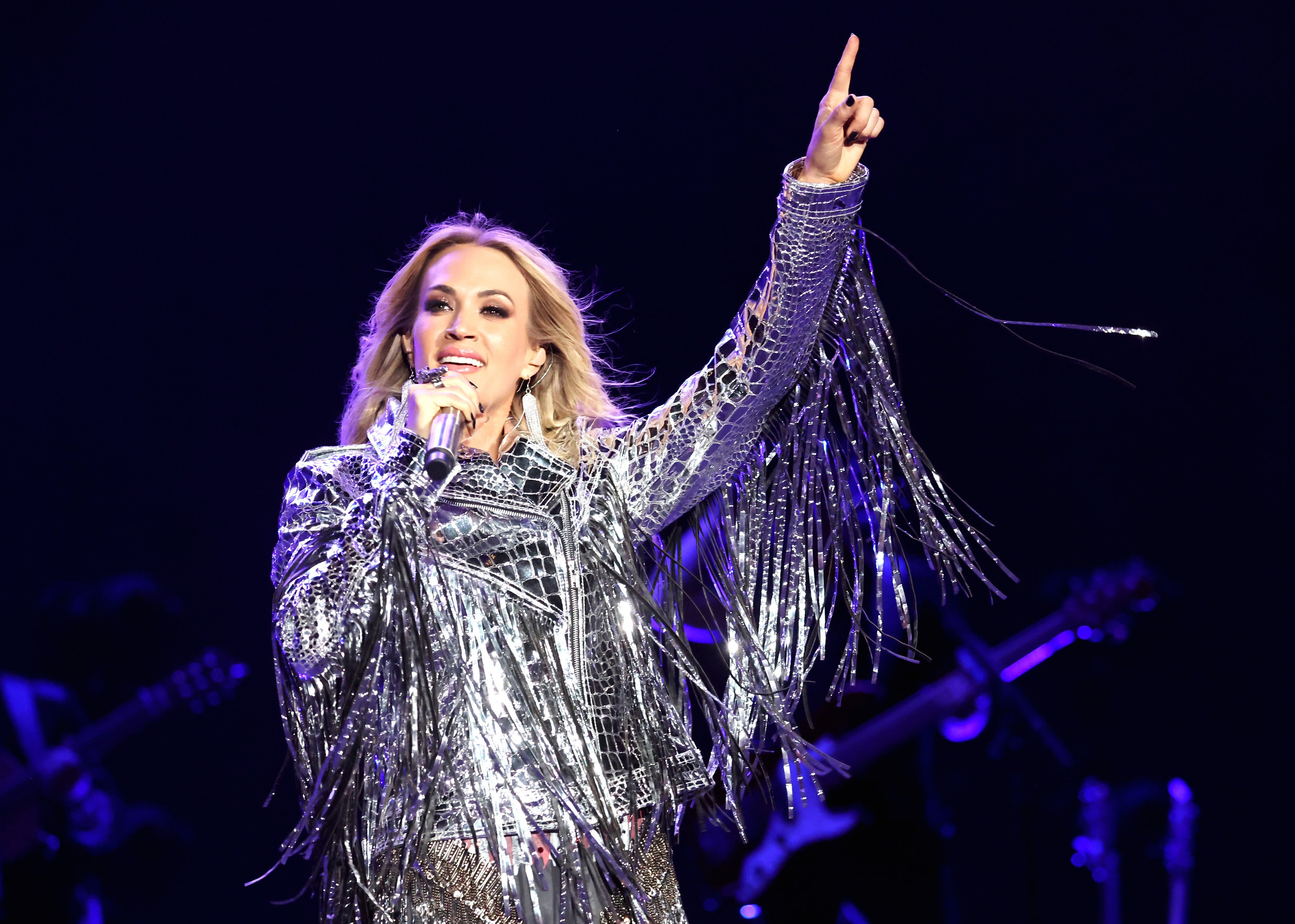 Carrie Underwood stands on stage and points at the sky while wearing a metallic outfit