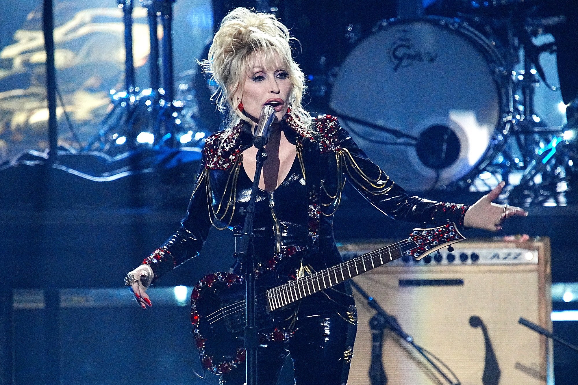 Dolly Parton performs on stage with an electric guitar while wearing a black leather jumpsuit