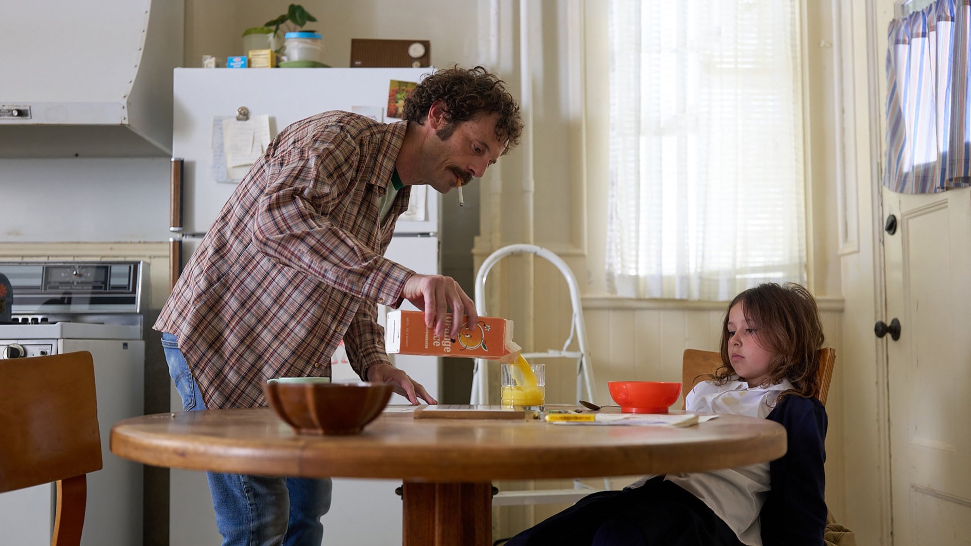 'Fairyland' Scoot McNairy as Steve Abbott and Nessa Dougherty as Younger Alysia Abbott. Steve is pouring orange juice into a glass with Alysia sitting at the table.