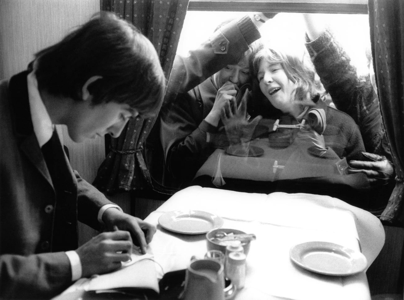 A black and white picture of George Harrison sitting at a table while fans look at him through the window.