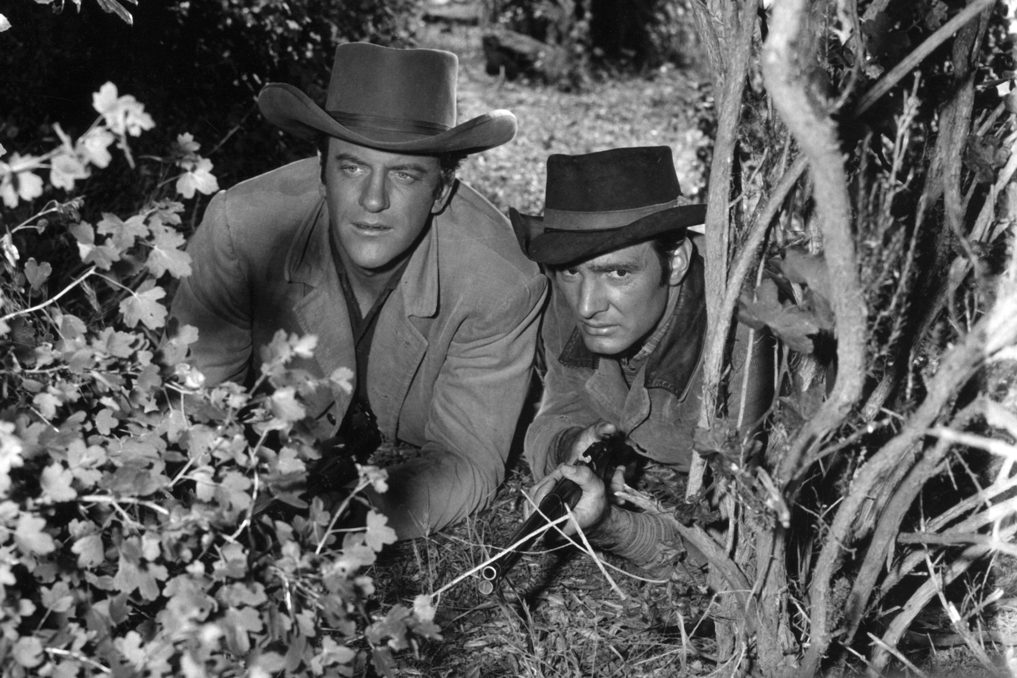 'Gunsmoke' James Arness as Marshal Matt Dillon and Dennis Weaver as Chester Goode crouched down in bushes wearing Western costumes. Chester is holding a gun.