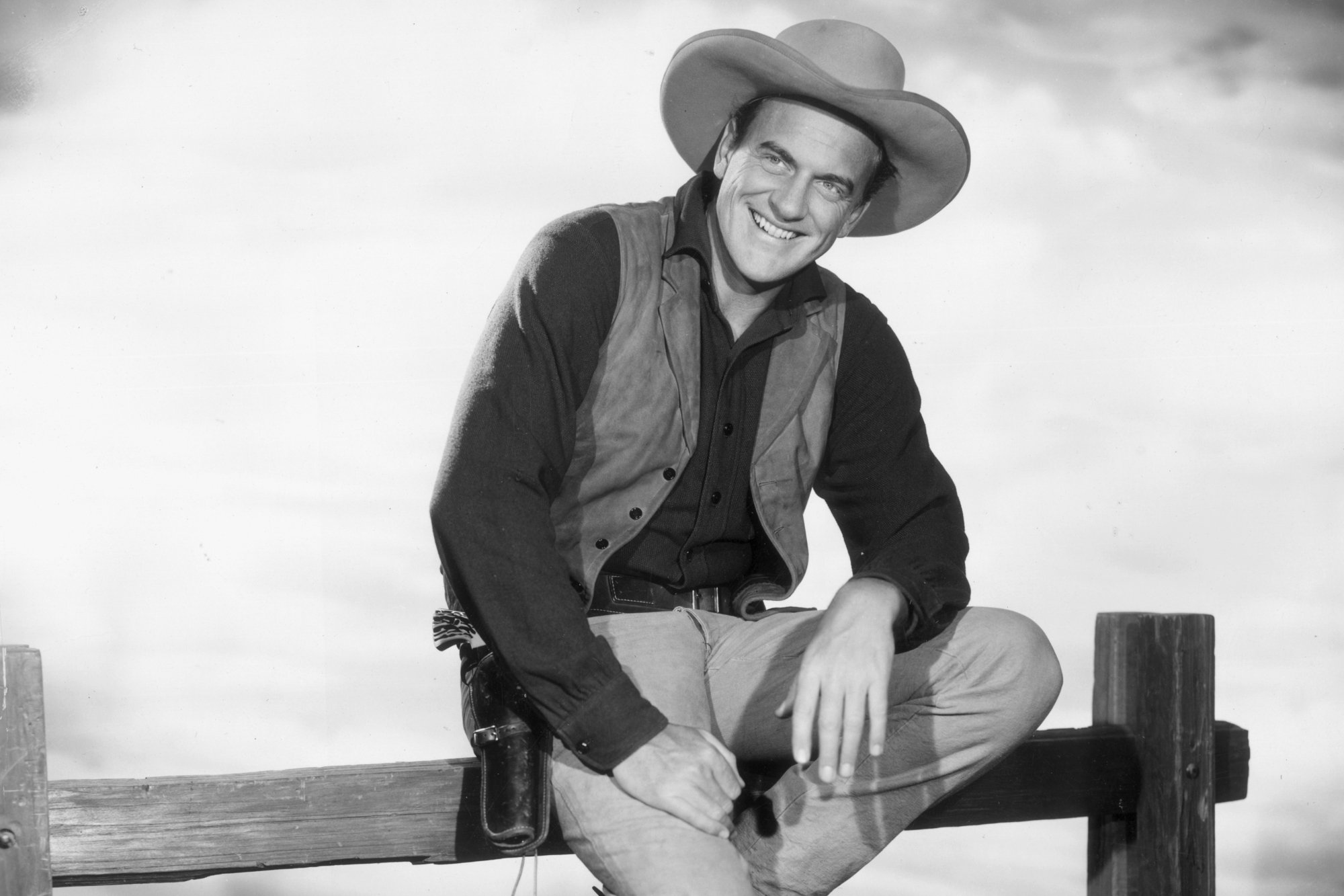 'Gunsmoke' James Arness as U.S. Marshal Matt Dillon in a black-and-white picture wearing his marshal uniform and sitting on a fence.