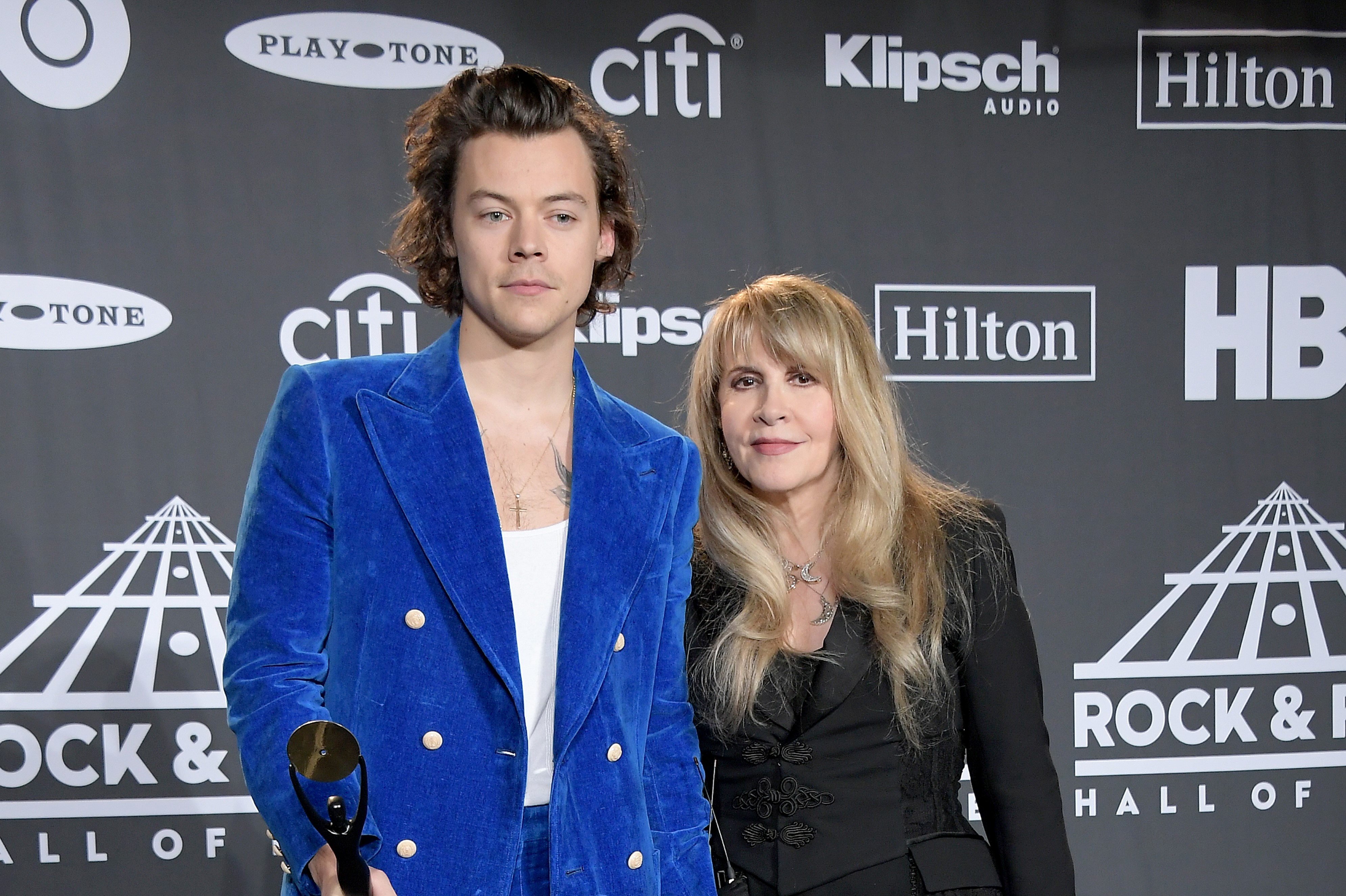 Harry Styles wears blue and Stevie Nicks wear black. They stand shoulder to shoulder.