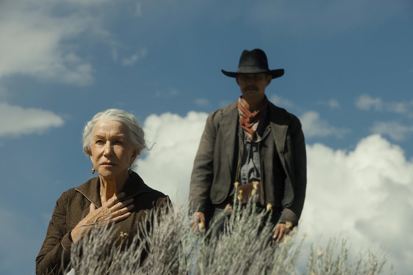 Helen Mirren as Cara Dutton kneeling down in tall grass with Zane Davis behind her in Taylor Sheridan's '1923'