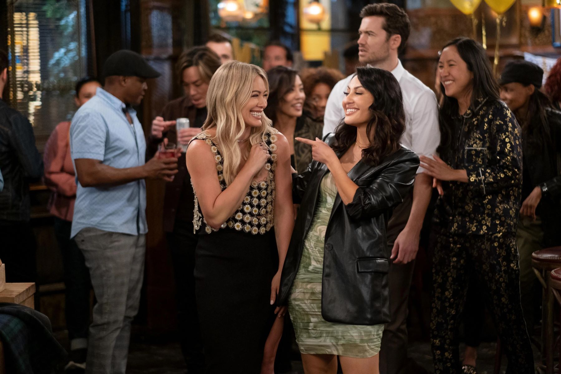 Hilary Duff and Francia Raisa, in character as Sophie and Valentina in 'How I Met Your Father' Season 2, share a scene at the bar. Sophie wears a daisy crop top over a black dress. Valentina wears a black leather jacket over a green and white dress.