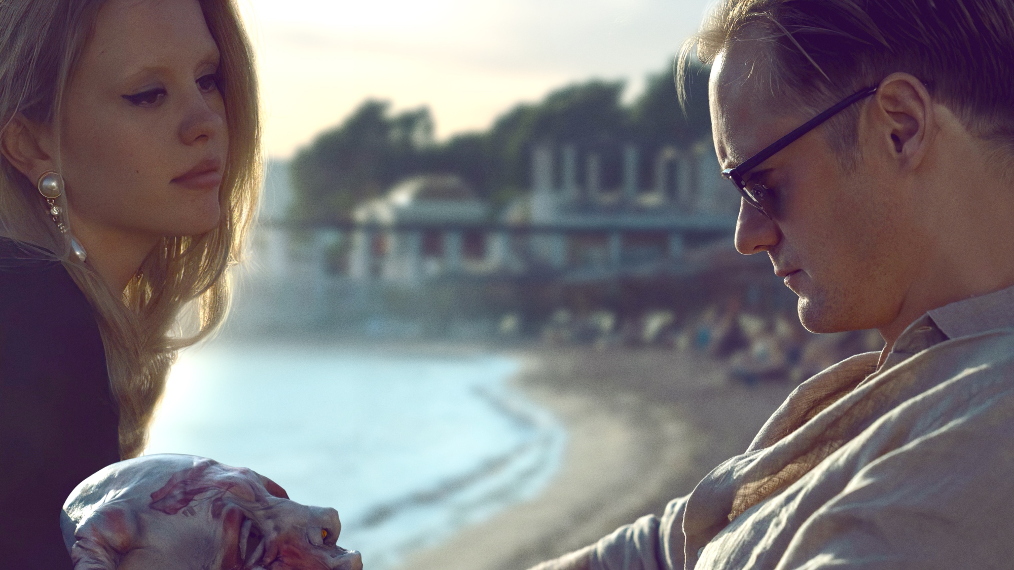 'Infinity Pool' Mia Goth as Gabi and Alexander Skarsgård as James Foster. James is looking down at a mask, while Gabi looks at James in front of a body of water.