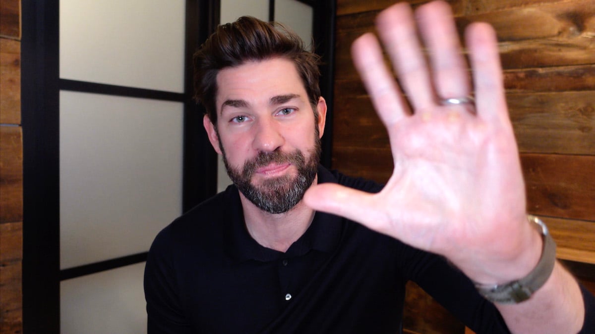 John Krasinski waves to the camera while speaking using video conferencing software.