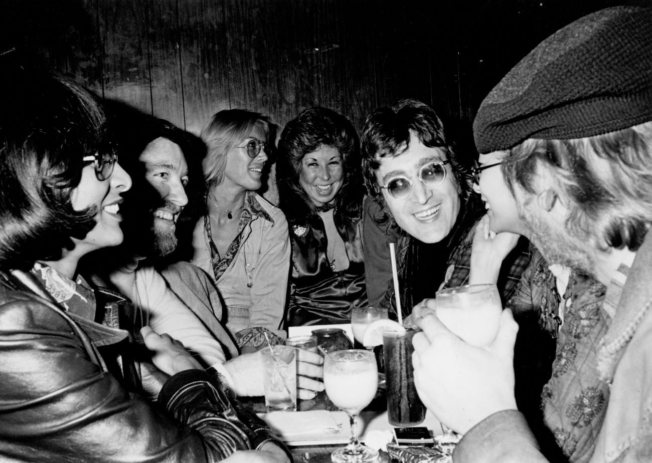 A black and white picture of John Lennon wearing sunglasses and sitting around a crowded table. 