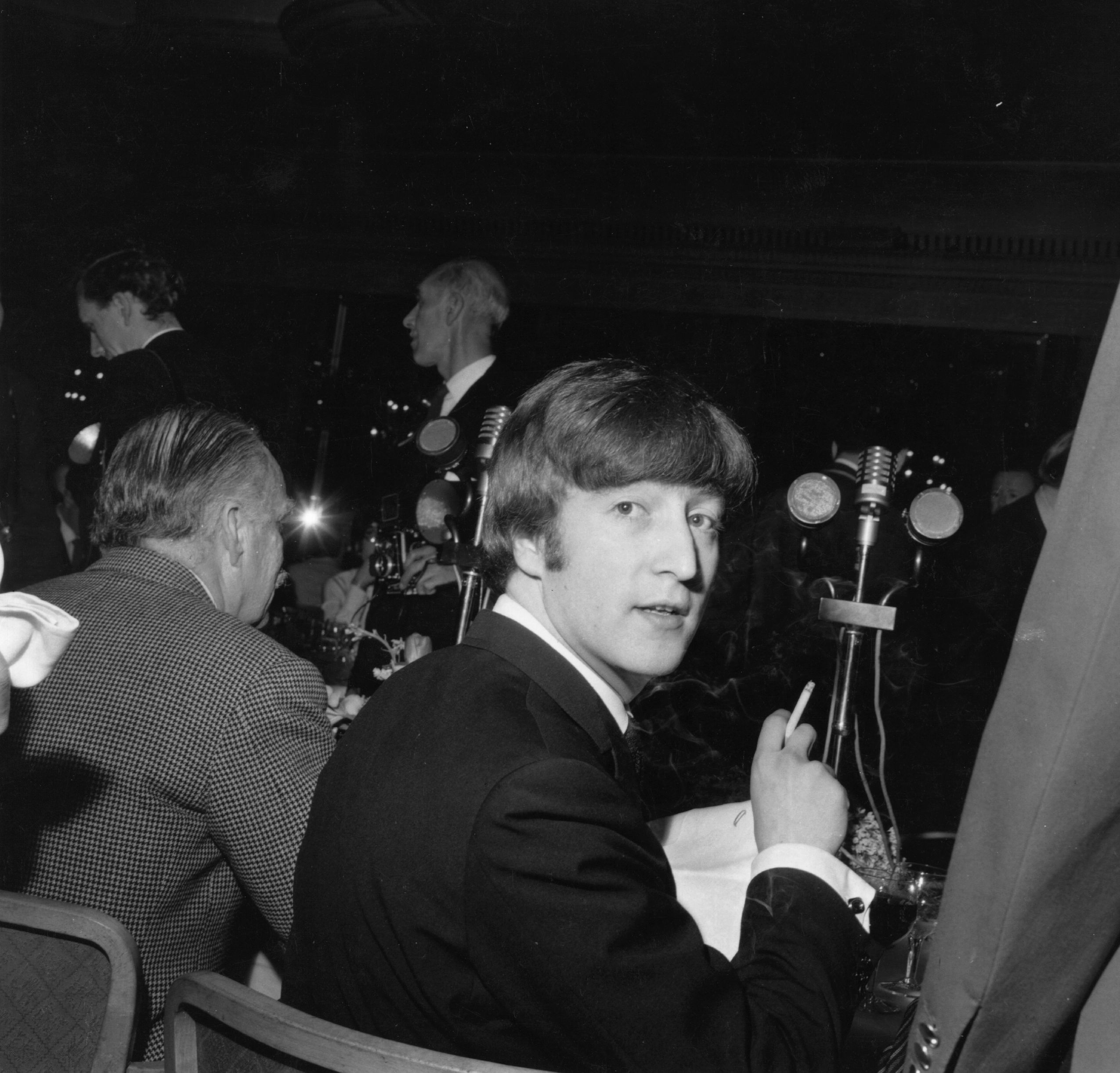 A black and white picture of John Lennon sitting at a table with a cigarette at a literary luncheon.