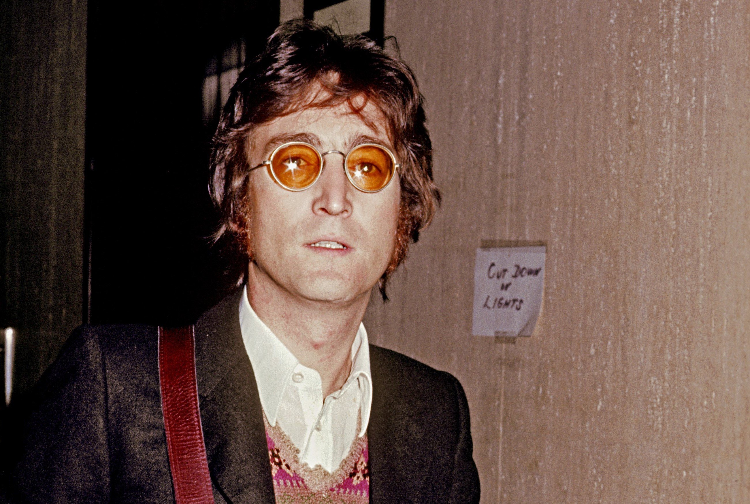 John Lennon wears sunglasses and carries a bag over his shoulder.
