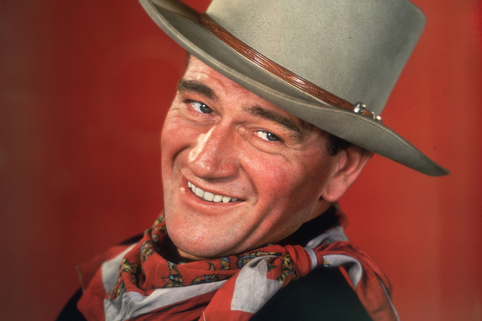 John Wayne, a star of Western movies. He's wearing a cowboy hat, smiling in front of a red background.