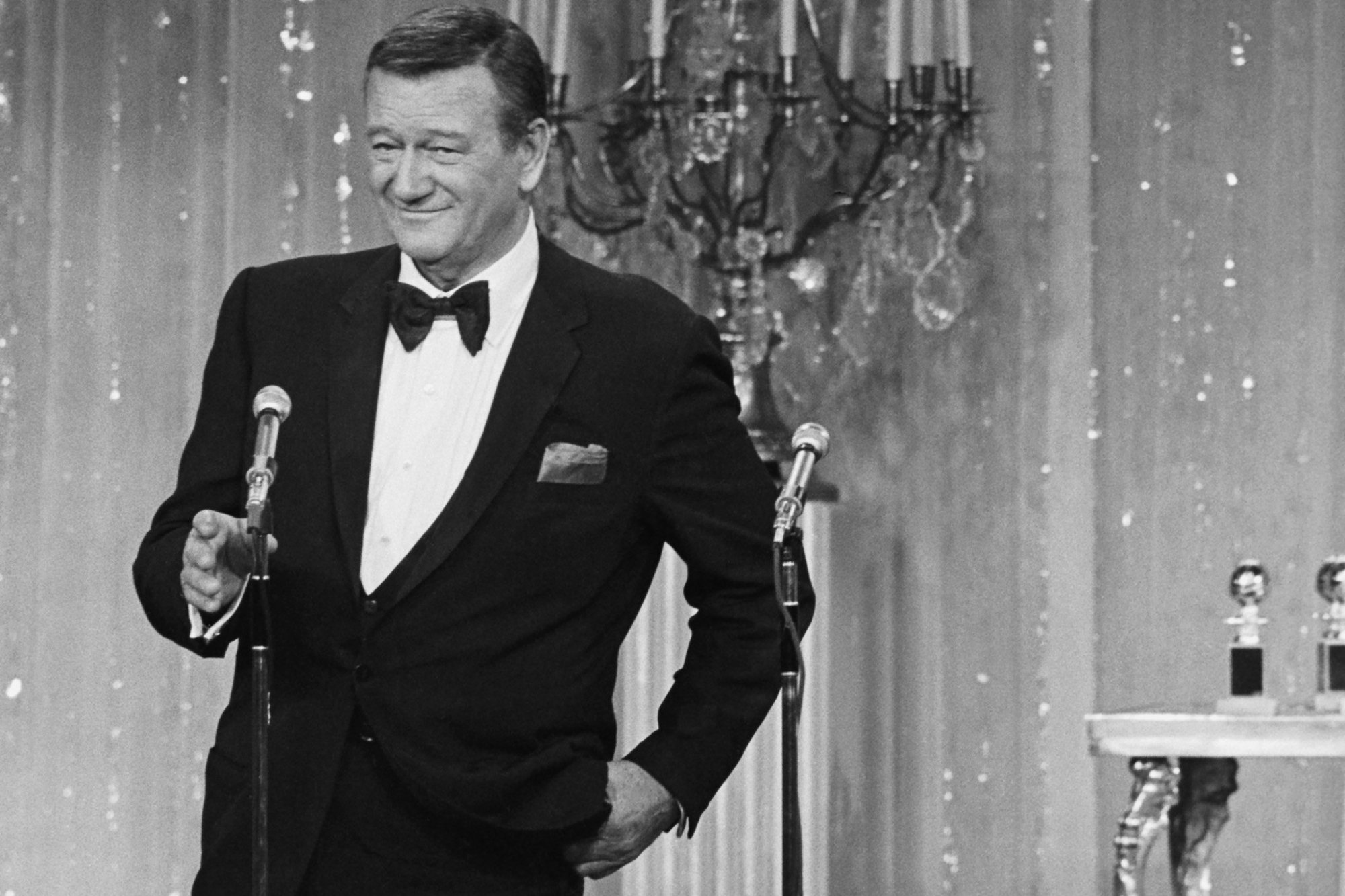 John Wayne at the Golden Globes in a black-and-white picture wearing a tux, holding onto the microphone
