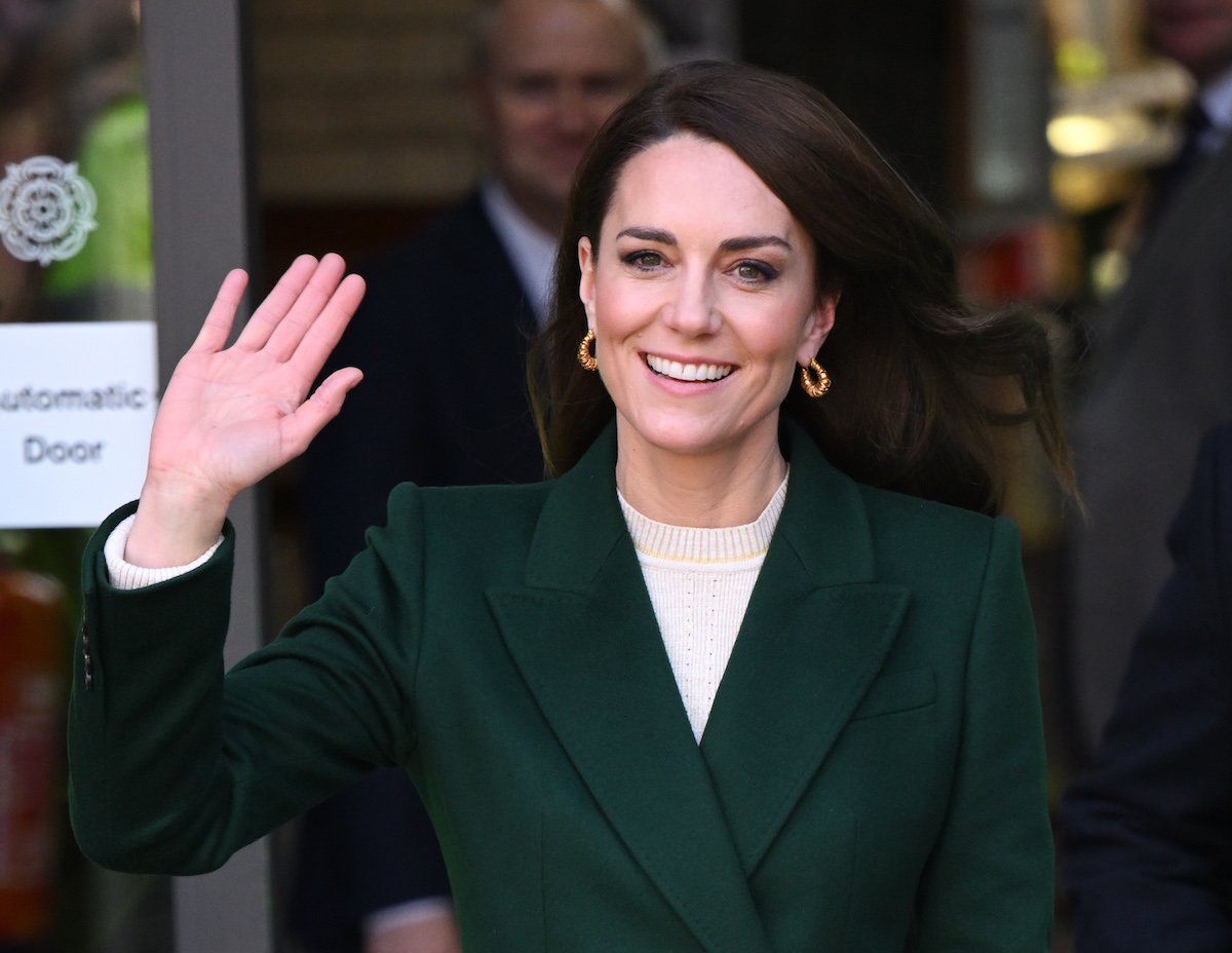 Kate Middleton smiles and waves, wearing a suit and gold earrings.
