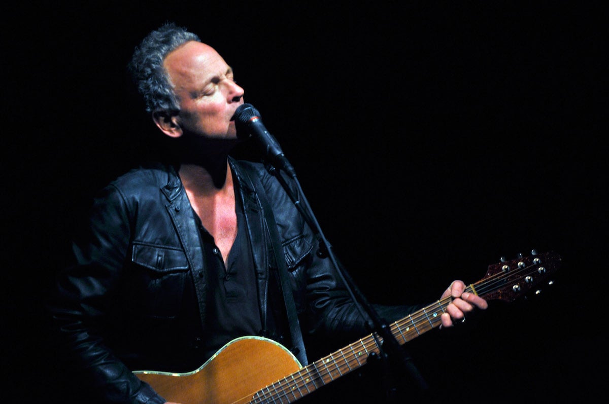 Fleetwood Mac guitarist Lindsey Buckingham performs on stage, singing into a microphone and playing guitar with his eyes closed.