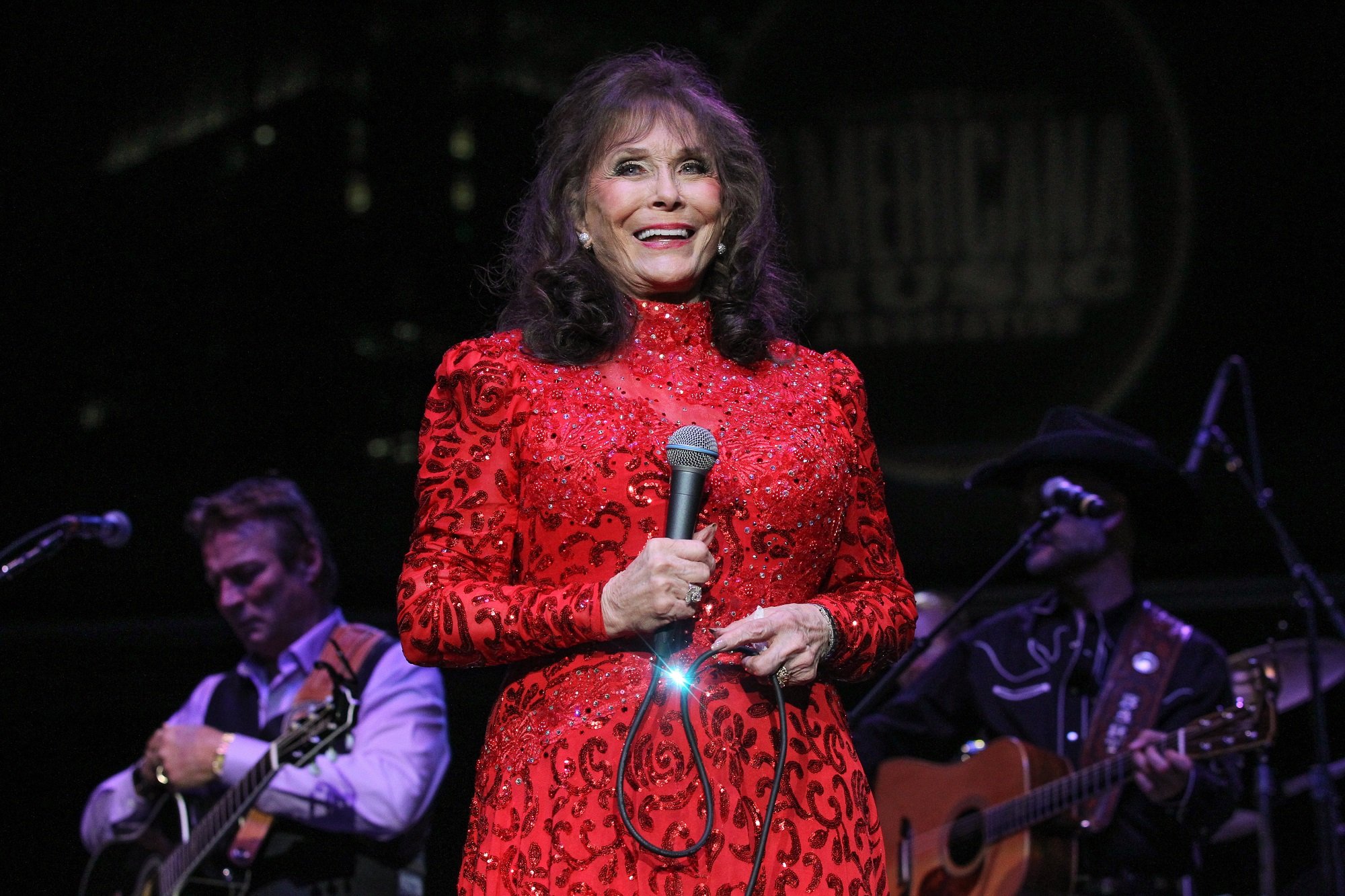 Loretta Lynn smiles in a bright red gown while holding a microphone