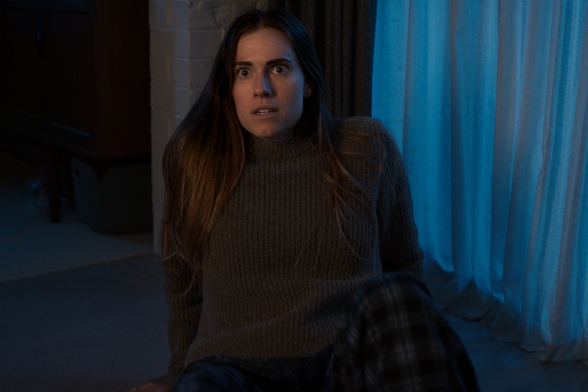 'M3GAN' Allison Williams as Gemma sitting on the ground in front of curtains, looking terrified