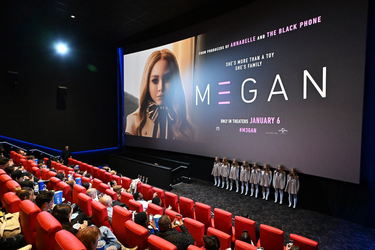 A movie theater is filled with audience members, looking at a screen with the "M3GAN" logo on it while models dressed as the title character pose.
