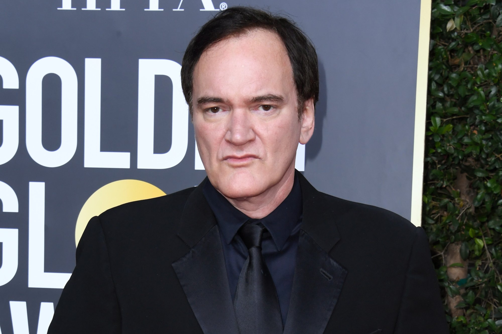 Movie director and screenwriter Quentin Tarantino. He has a plain expression on his face, wearing a black suit, standing in front of a Golden Globes sign.