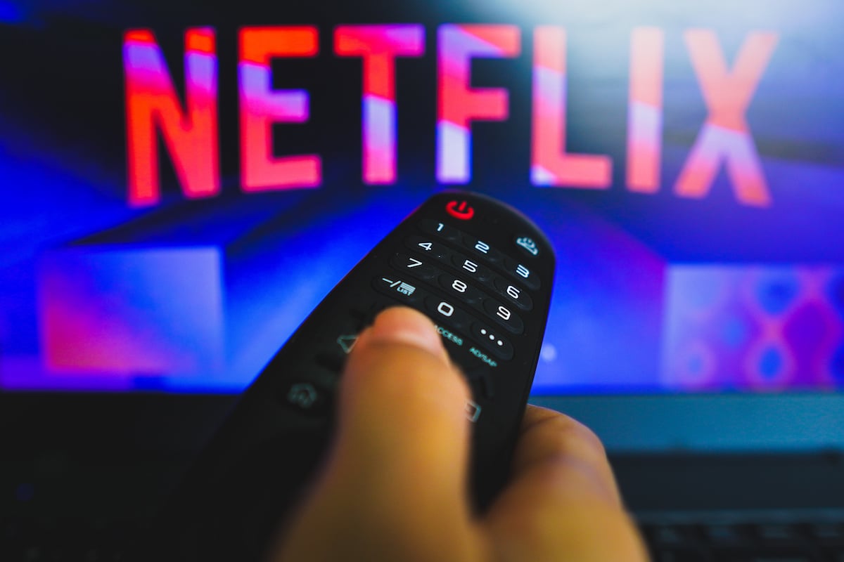A hand holding a TV remote in front of a TV showing the Netflix logo.