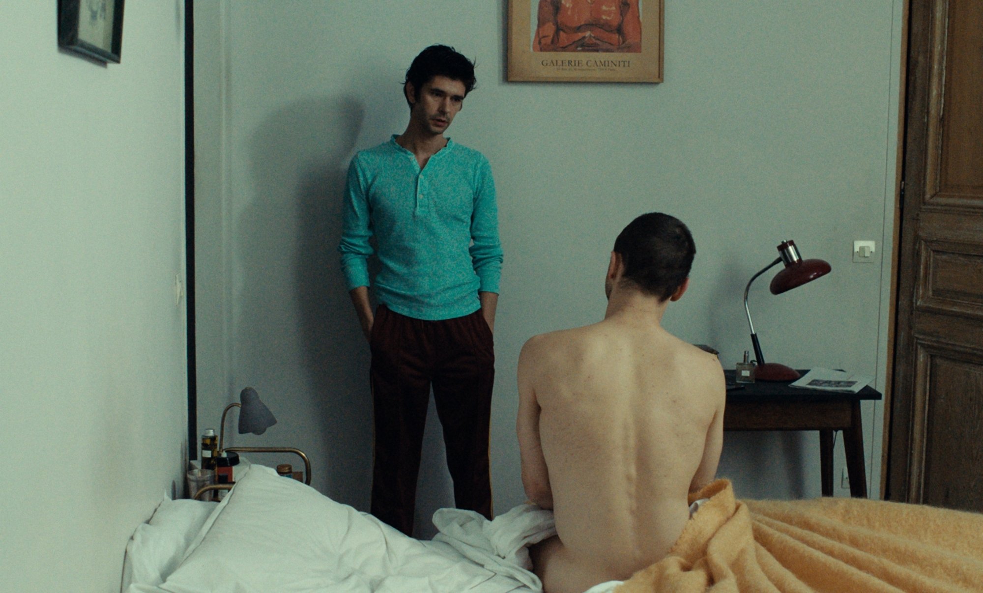'Passages' Ben Whishaw as Martin and Franz Rogowski as Tomas. Martin is standing with his back to the wall in a bedroom. Tomas is sitting naked on the bed looking at Martin.