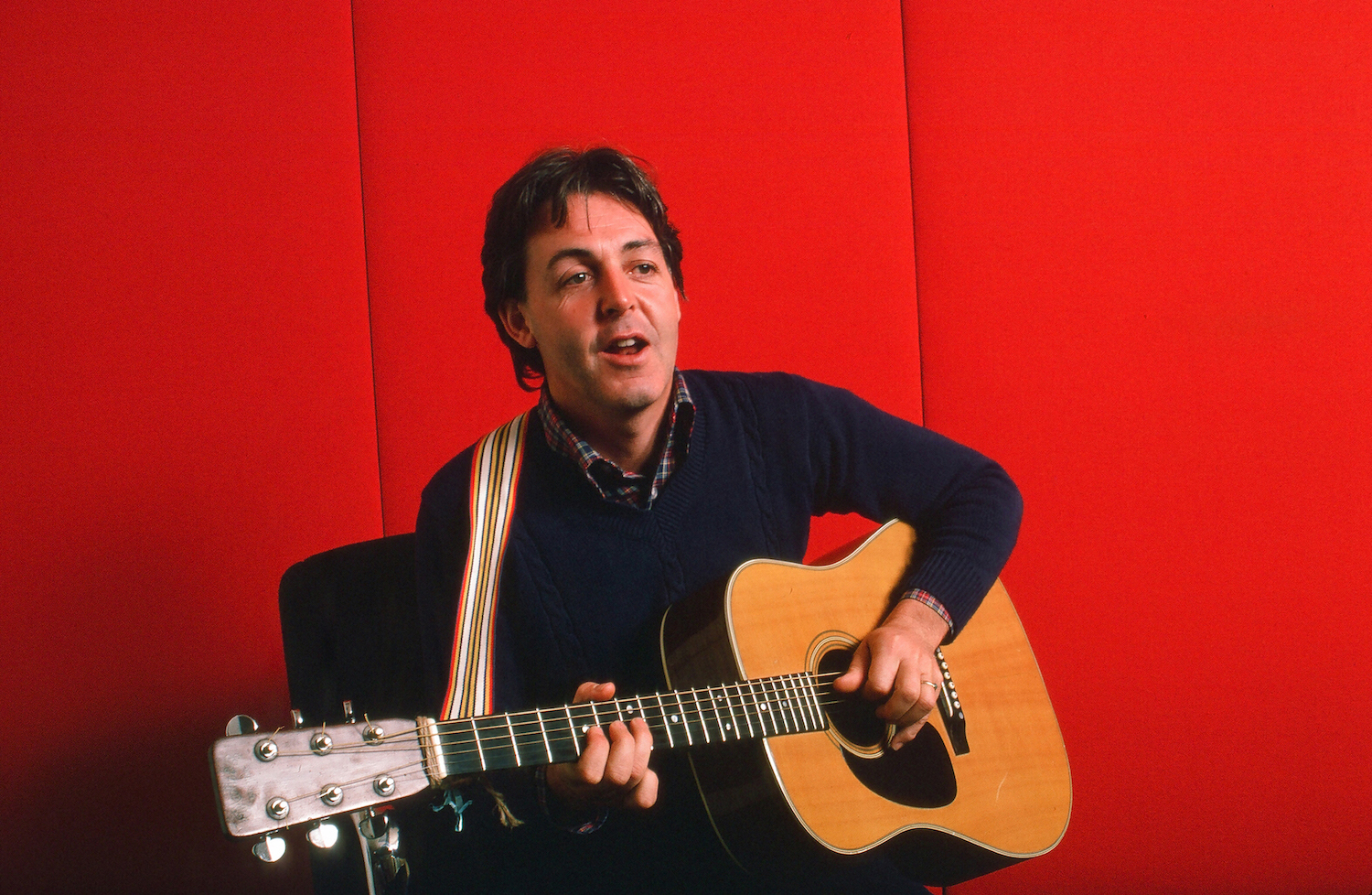 Portrait of British musician Paul McCartney as he plays acoustic guitar against a red background