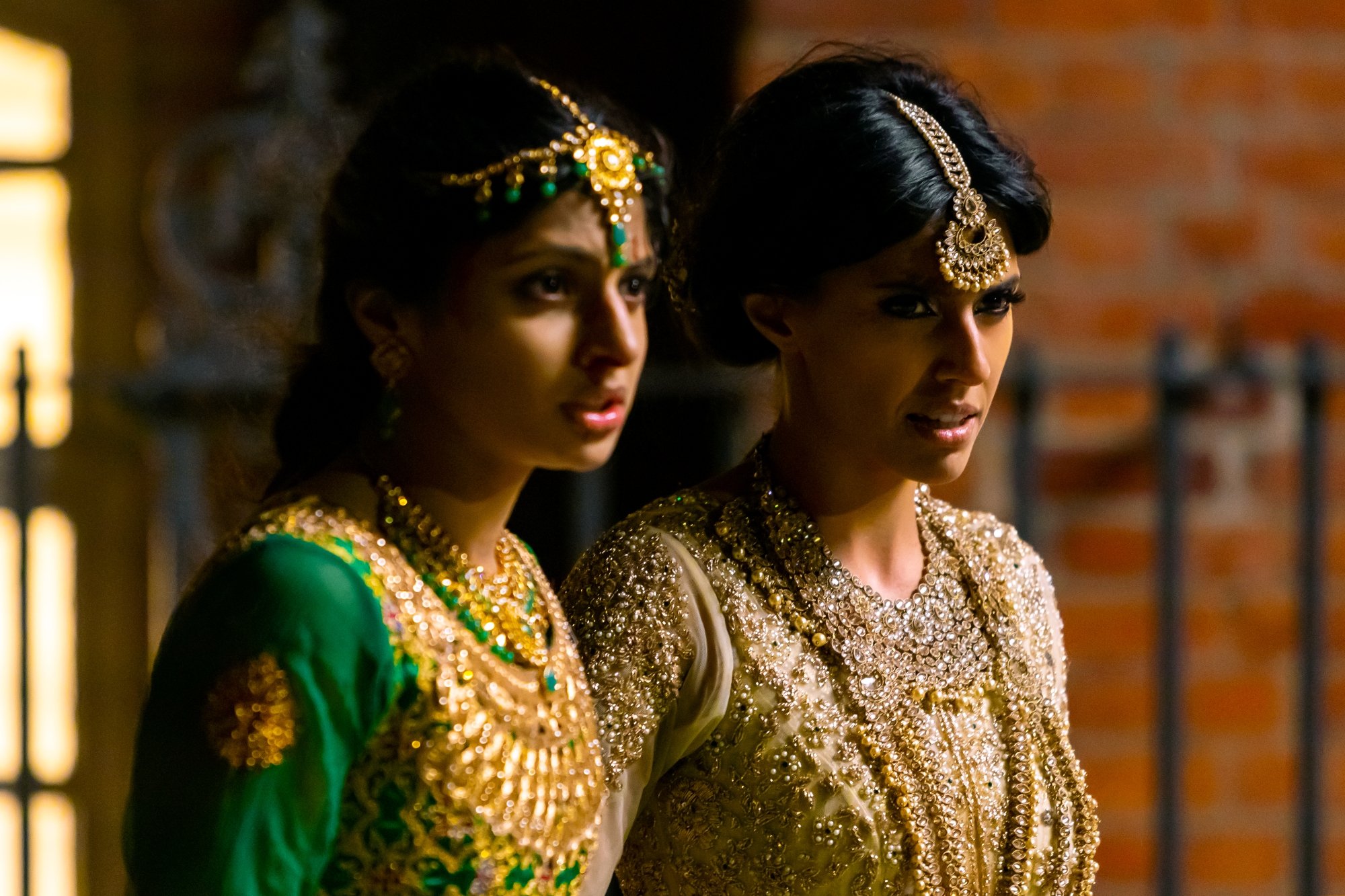 'Polite Society' Priya Kansara as Ria Khan and Ritu Arya as Lena looking shocked. Ria wearing a green and gold dress. Lena wearing a white and gold dress.