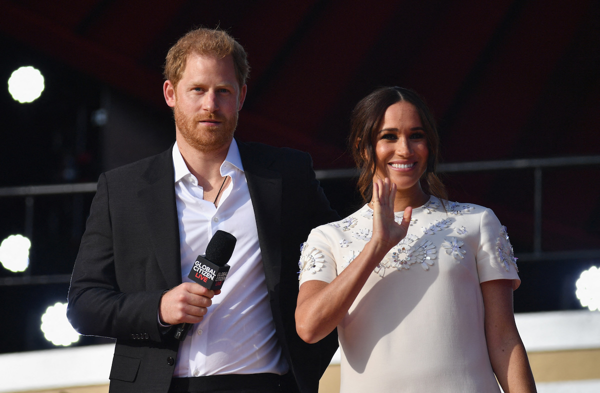 Prince Harry holds a microphone while Meghan Markle stands by his side and waves.