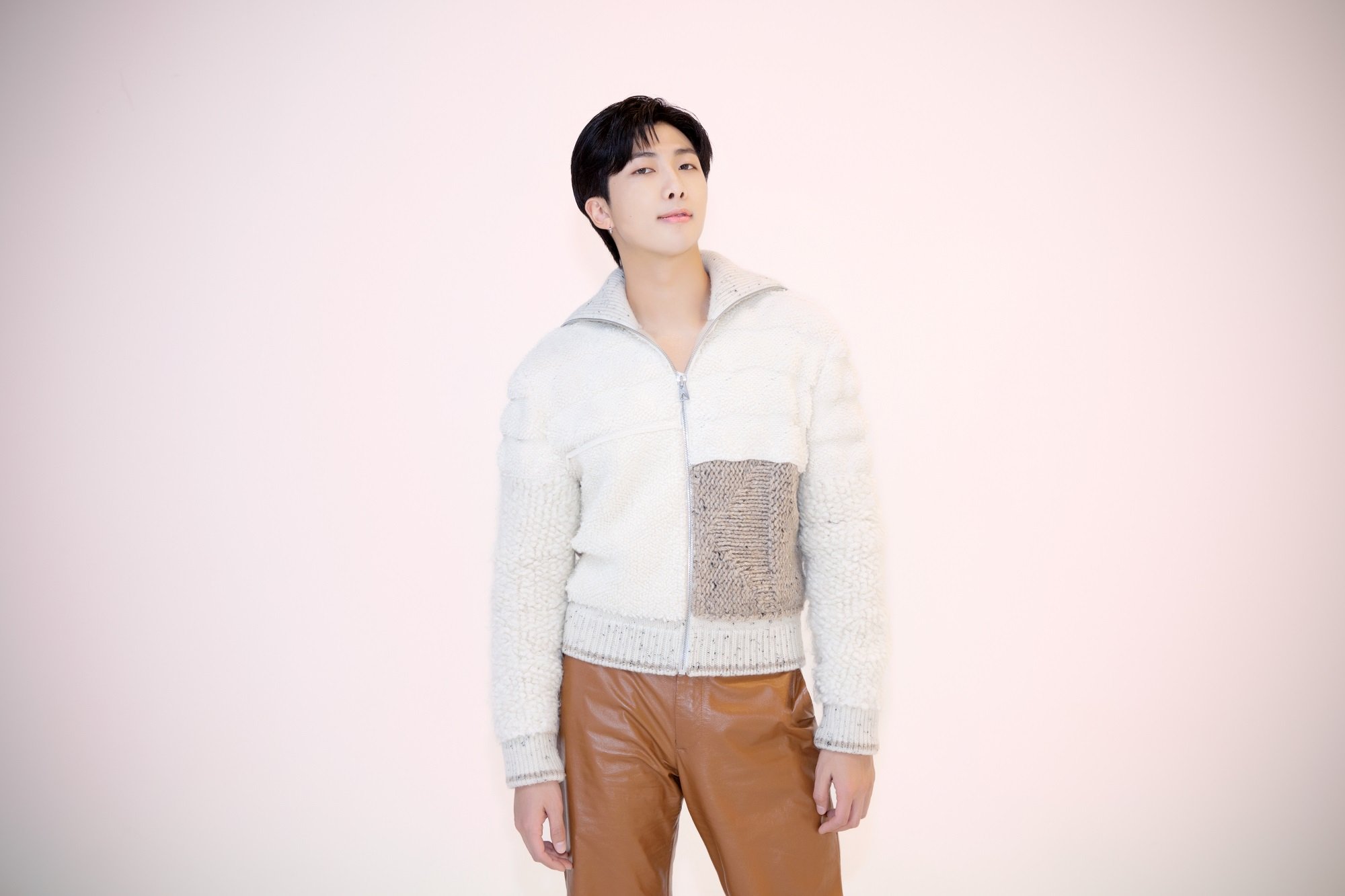 RM of BTS wears a white sweater and brown leather pants in front of a light backdrop