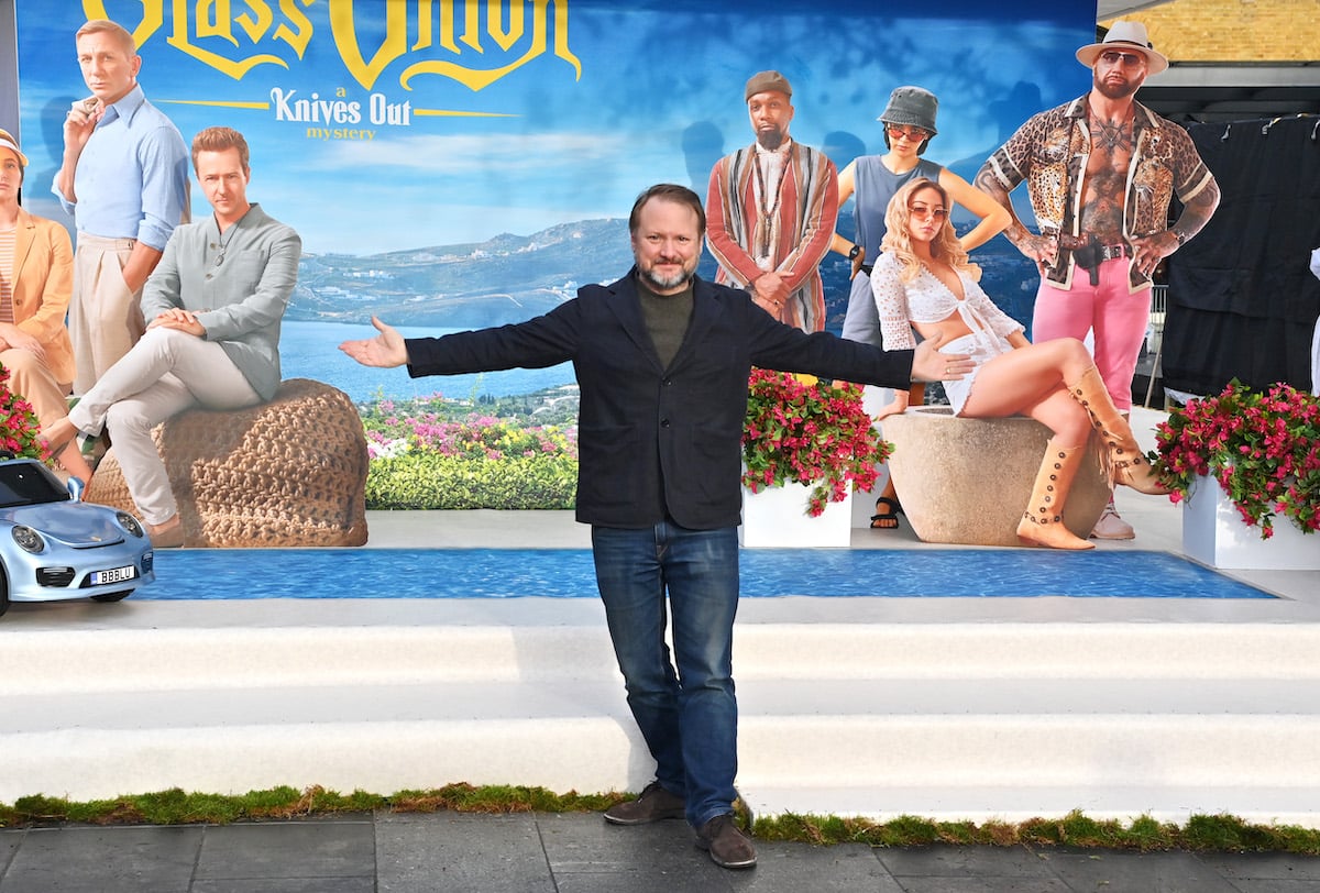 Director Rian Johnson poses with his arms outstretched in front of an oversized image of the "Glass Onion" movie poster.