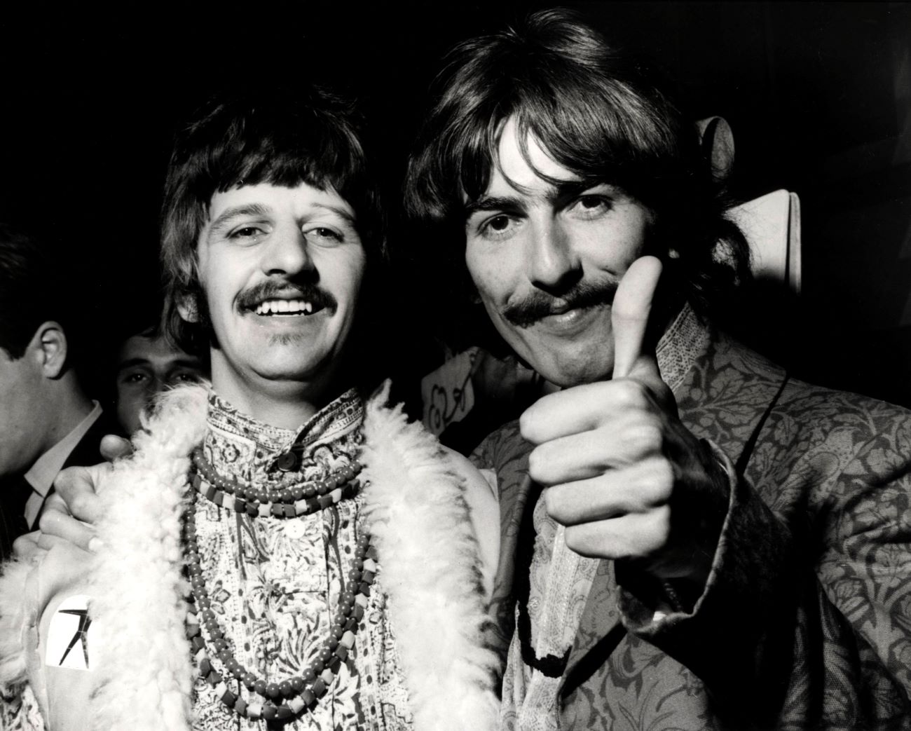 A black and white picture of George Harrison holding up a thumbs up with his arm around Ringo Starr's shoulders.