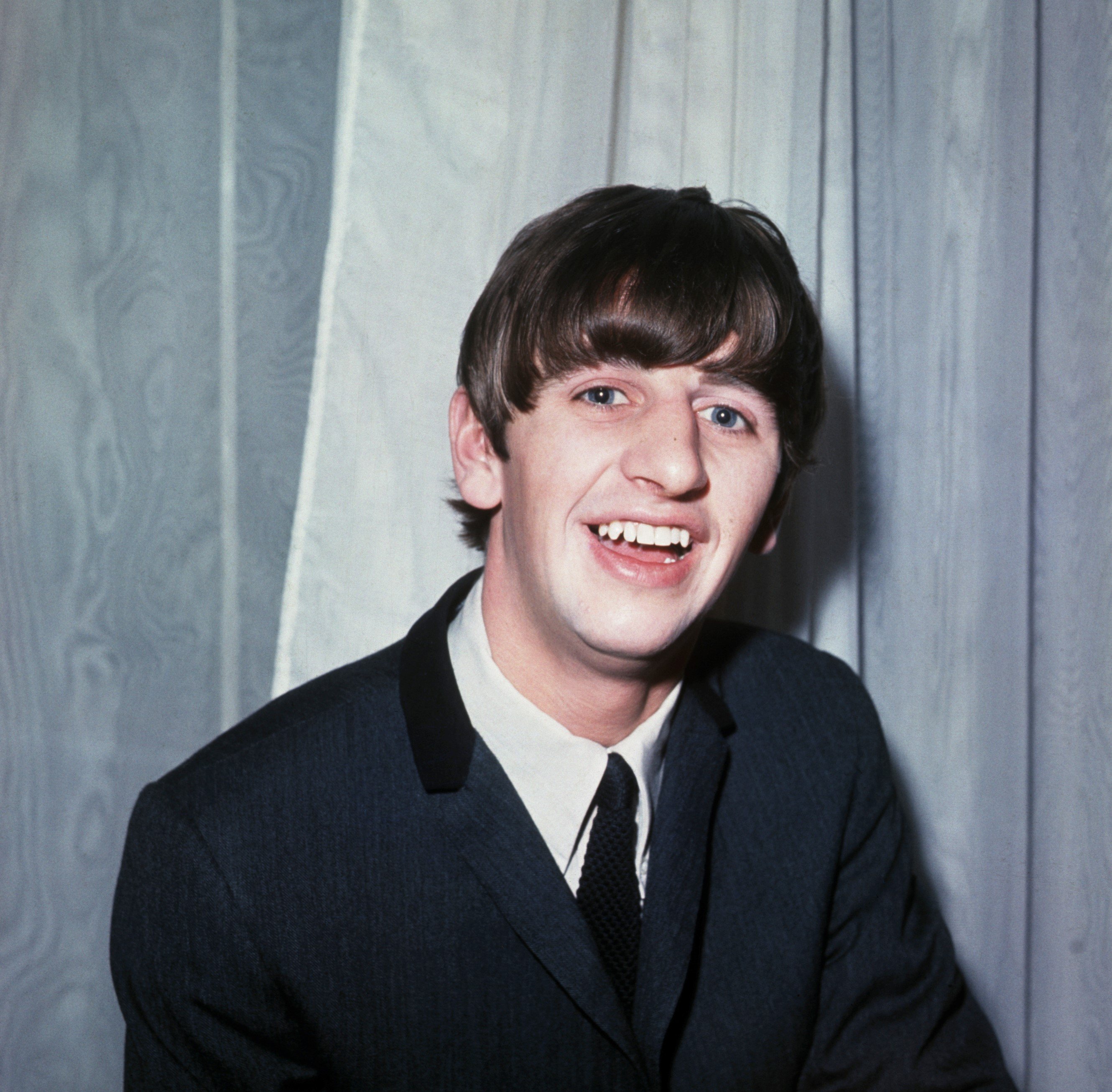 Ringo Starr wears a suit and poses in front of a gray wooden background.