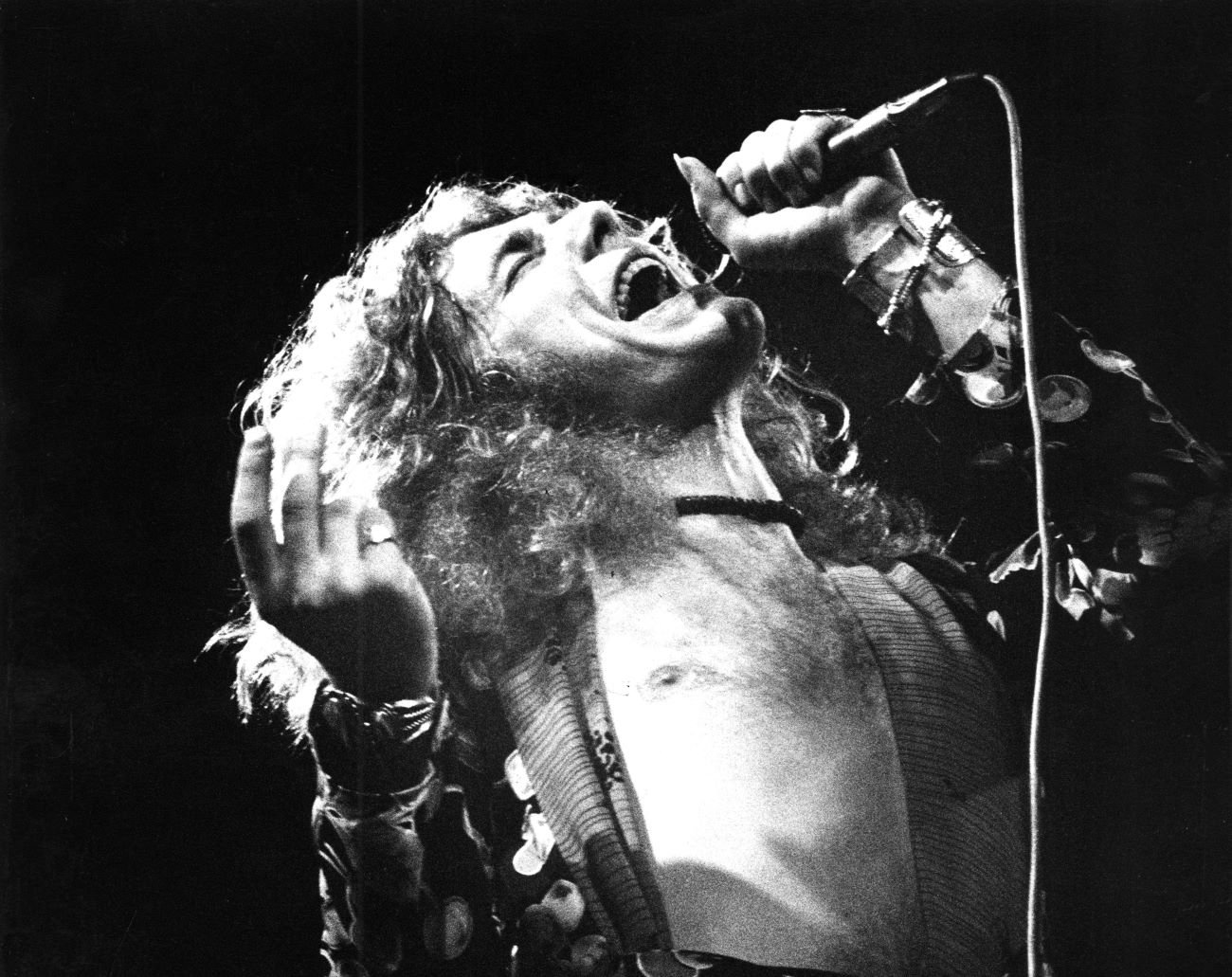 A black and white picture of Robert Plant singing into a microphone with his shirt open.