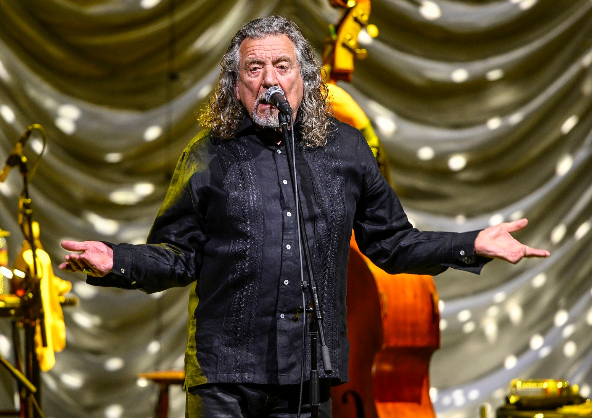 Robert Plant speaks into a microphone on stage with his arms held out