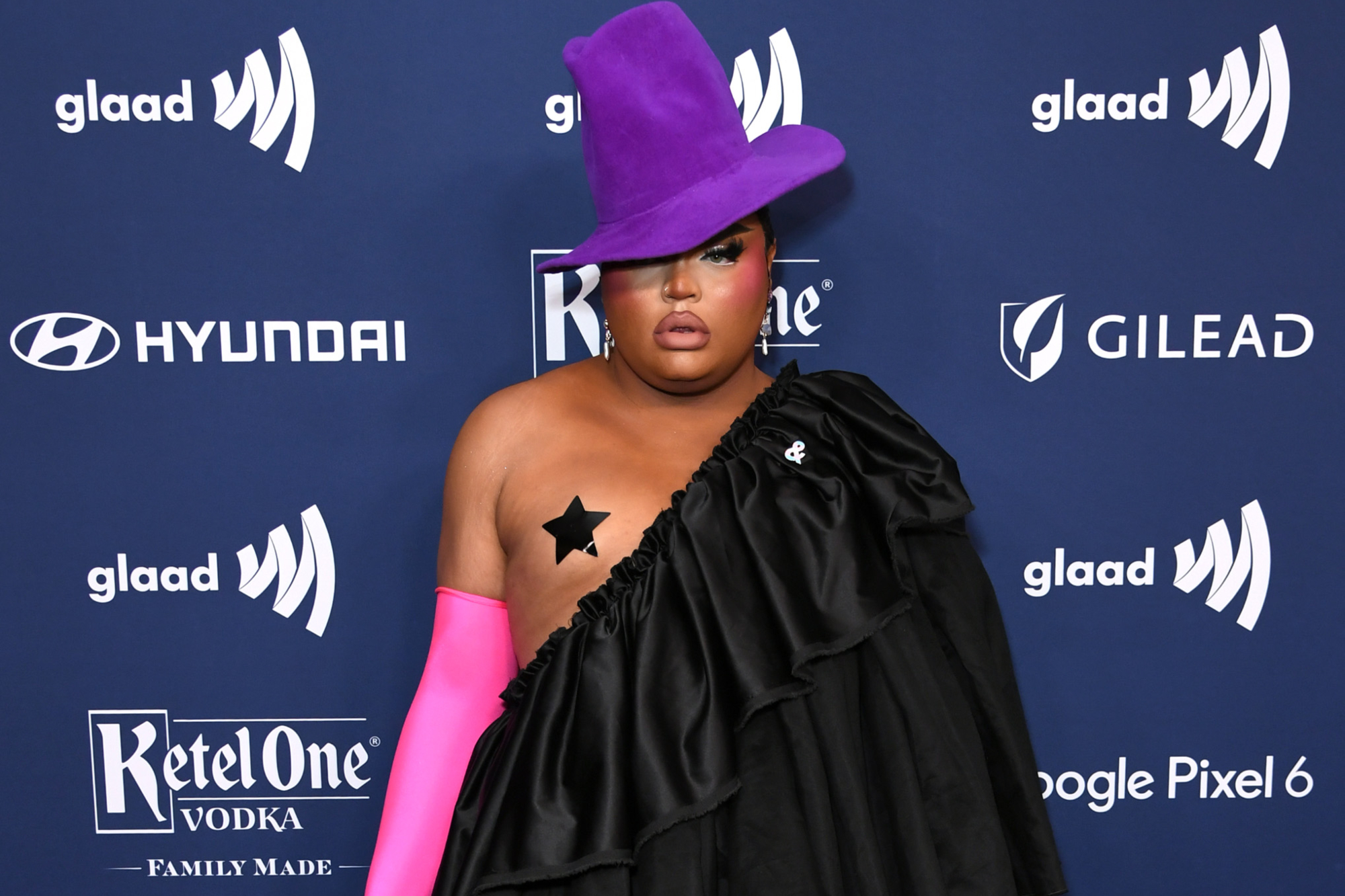 'RuPaul's Drag Race' Season 13 Kandy Muse wearing a purple hat, pink globes, and a black costume standing in front of a blue step and repeat