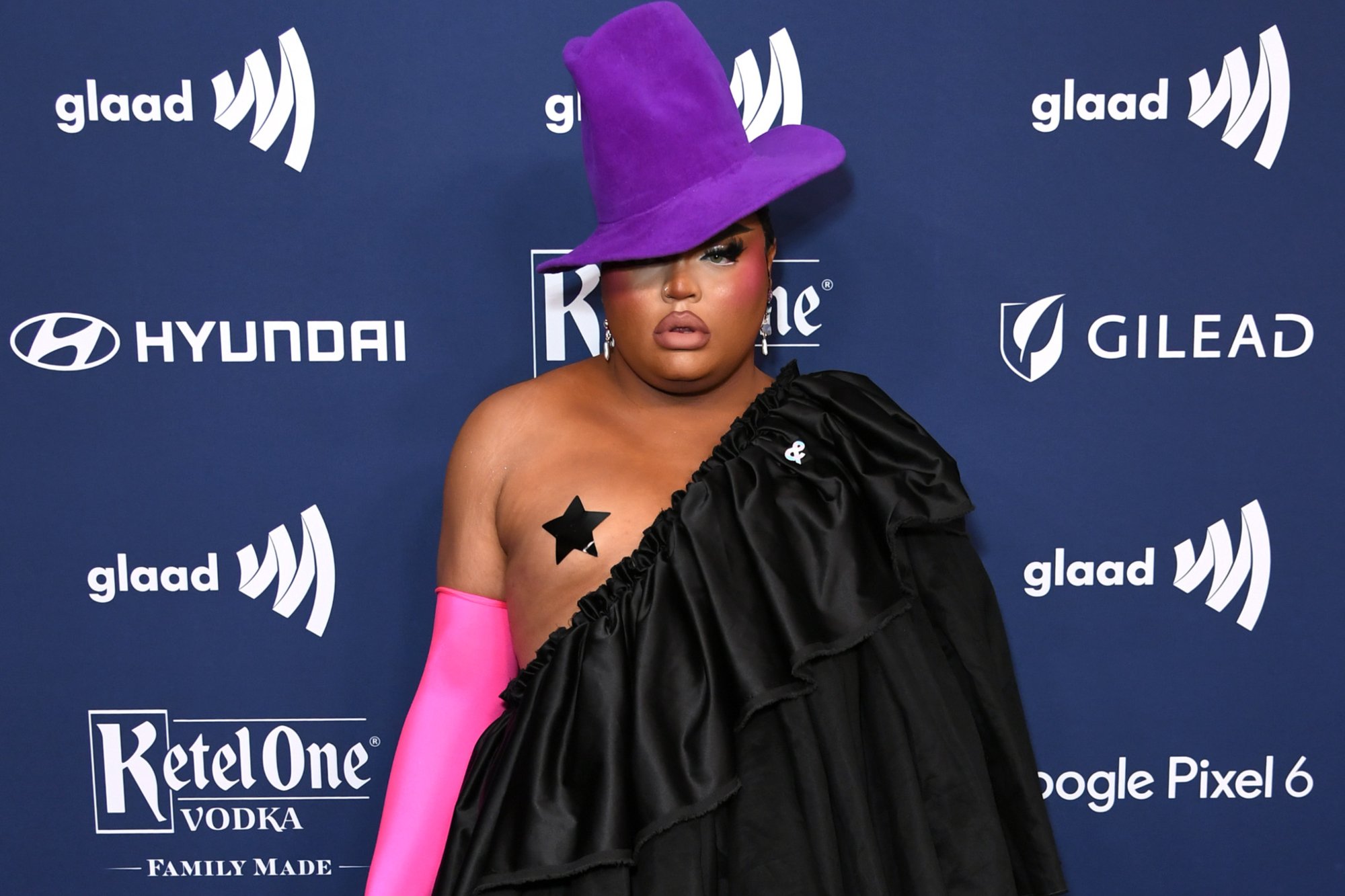 'RuPaul's Drag Race' Season 13 Kandy Muse wearing a purple hat, pink globes, and a black costume standing in front of a blue step and repeat