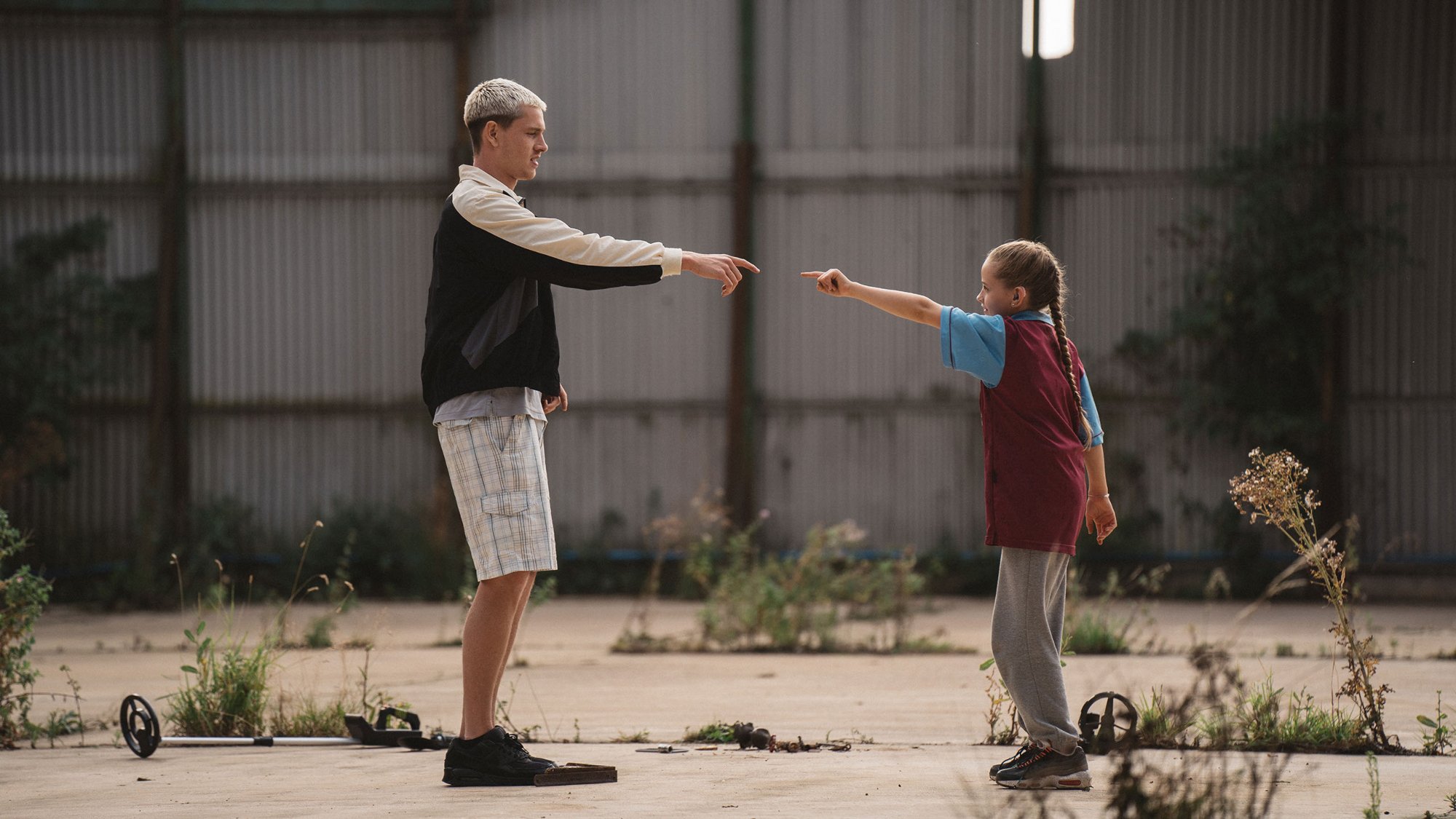 'Scrapper' Harris Dickinson as Jason and Lola Campbell as Georgie pointing at each other, standing on asphalt in front of a metal gate