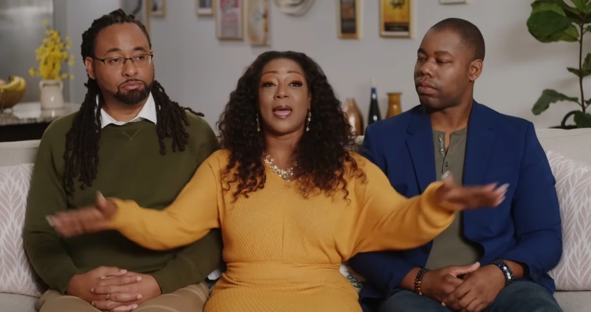 A woman wearing a yellow dress sits int he middle between her two husbands on a couch for an interview for 'Seeking Brother Husband' on TLC.