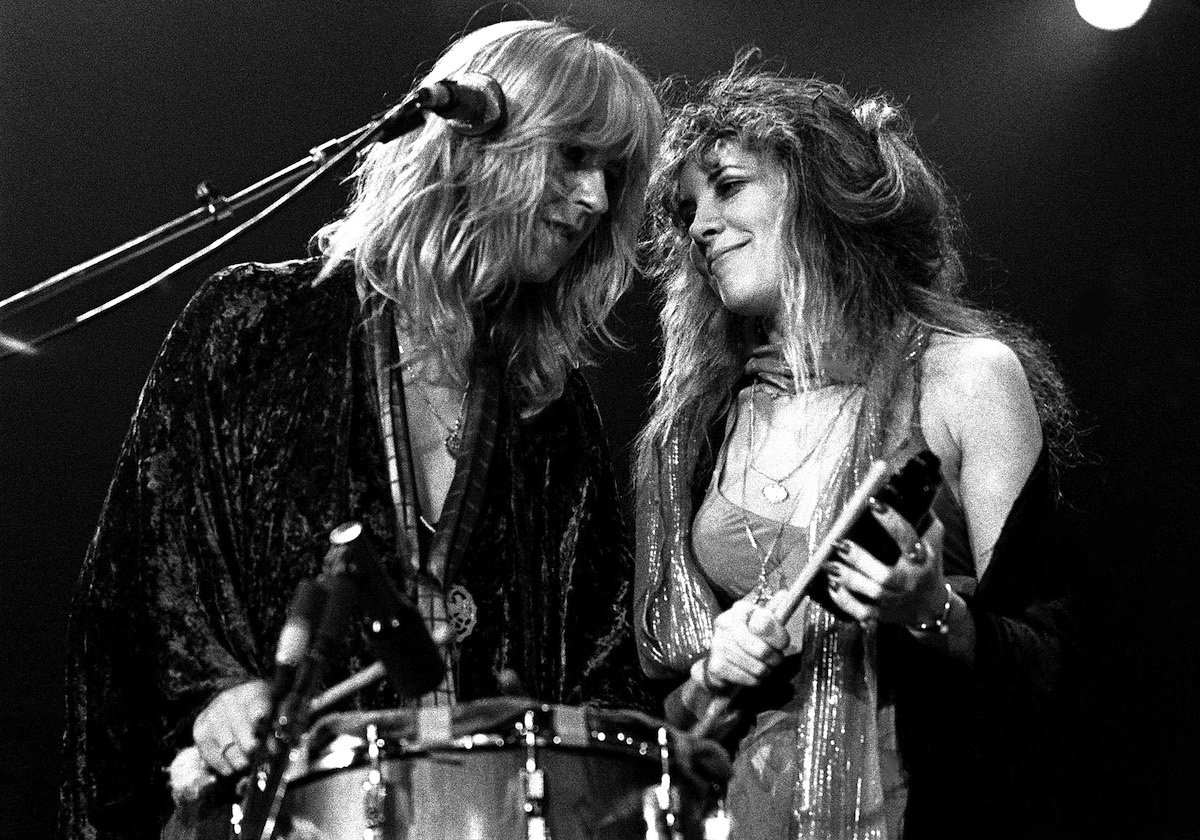 A black and white photo of Fleetwood Mac stars Christine McVie and Stevie Nicks performing on stage together, smiling and looking at each other.