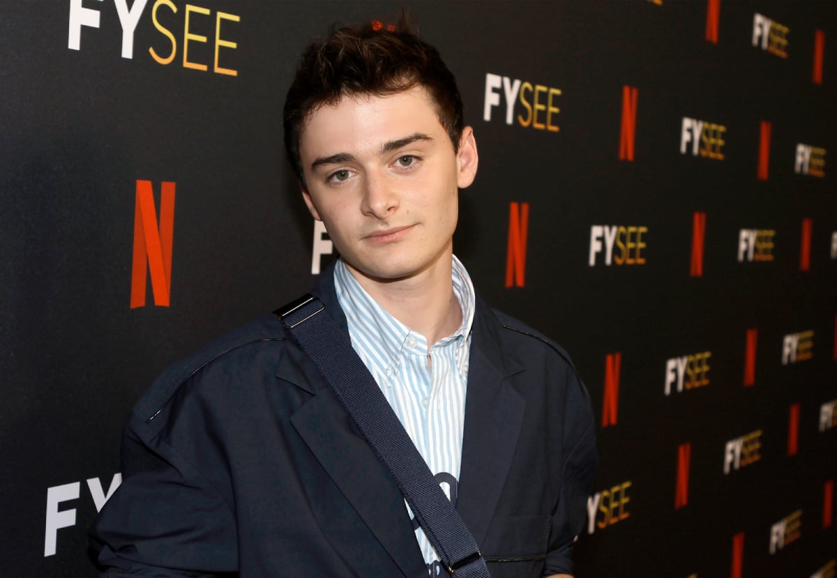 Stranger Things star Noah Schnapp wears a button-down shirt, black jacket, and cross-body bag.