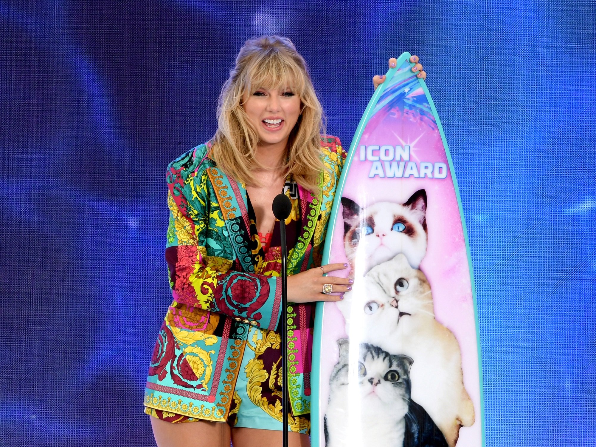Taylor Swift stands on stage next to a surfboard with photos of her cats Benjamin, Olivia, and Meredith