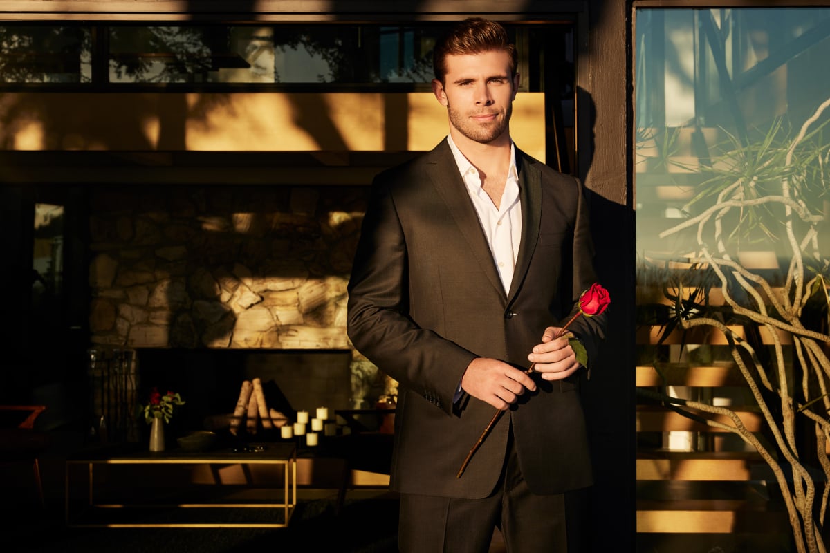 The Bachelor lead Zach Shallcross wears a black suit jacket and holds a rose.