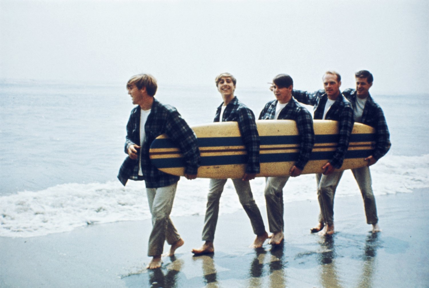 Rock and roll band The Beach Boys walk along the beach holding a surfboard (Dennis Wilson, David Marks, Mike Love, Carl Wilson, Brian Wilson)