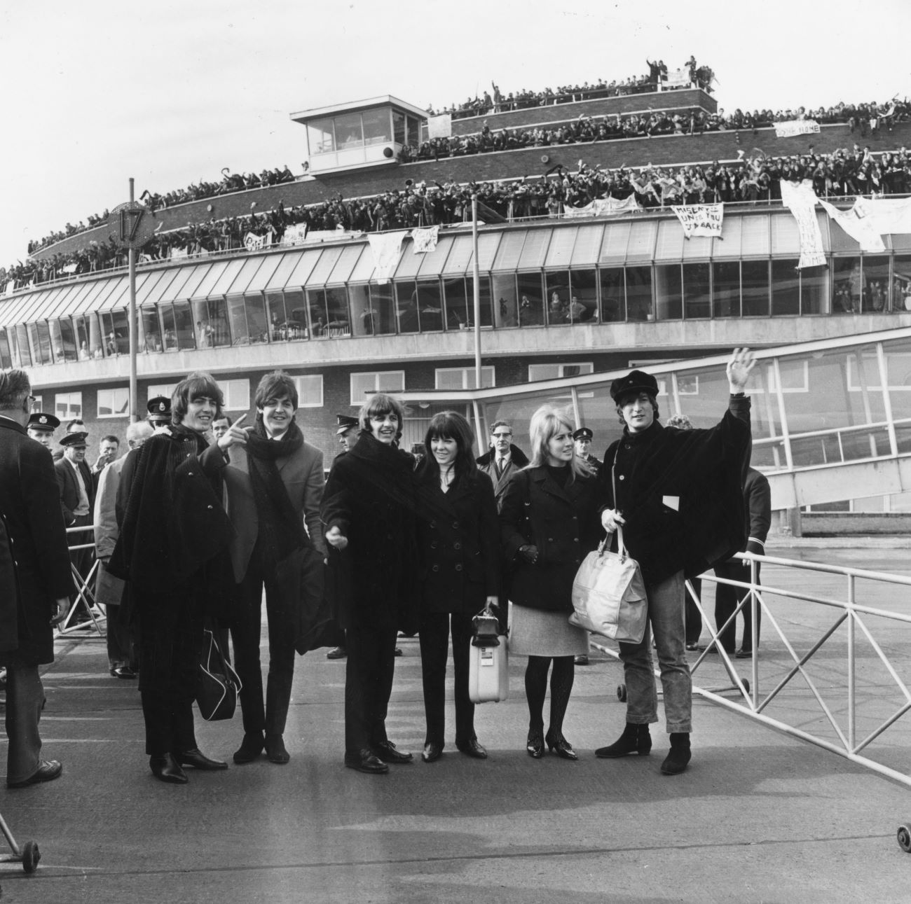 A black and white picture of George Harrison, Paul McCartney, Ringo Starr, Maureen Starkey, Cynthia Lennon, and John Lennon waving to fans at the airport.