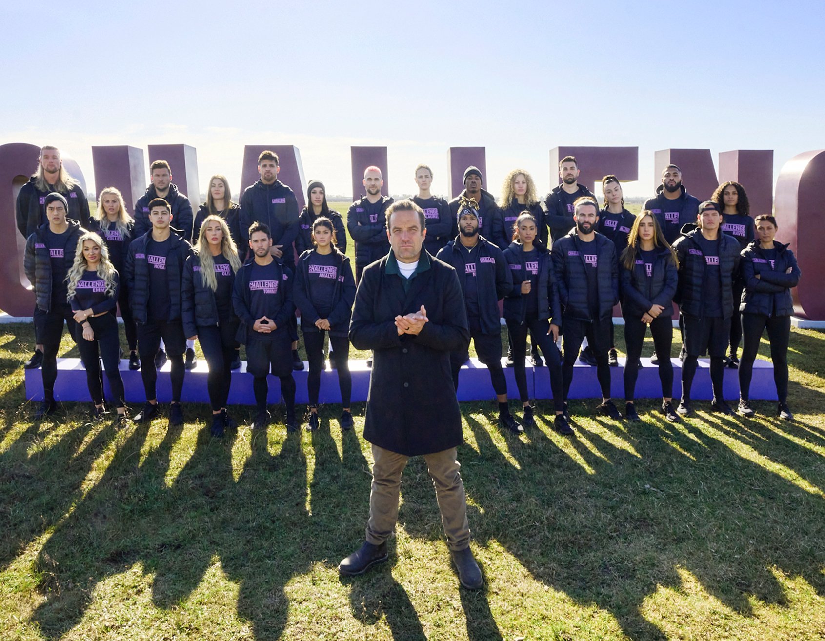 'The Challenge' Season 38 cast in uniforms standing behind T.J. Lavin on a field.