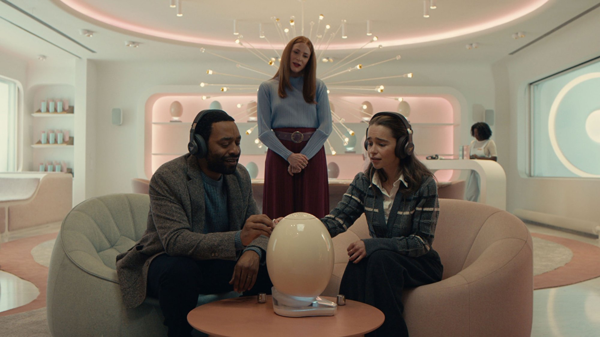 'The Pod Generation' Chiwetel Ejiofor as Alvy, Rosalie Craig as Linda Wozcheck, and Emilia Clarke as Rachel. Alvy and Rachel are sitting in front of the pod sitting on the table, touching it, while Linda is standing behind them.