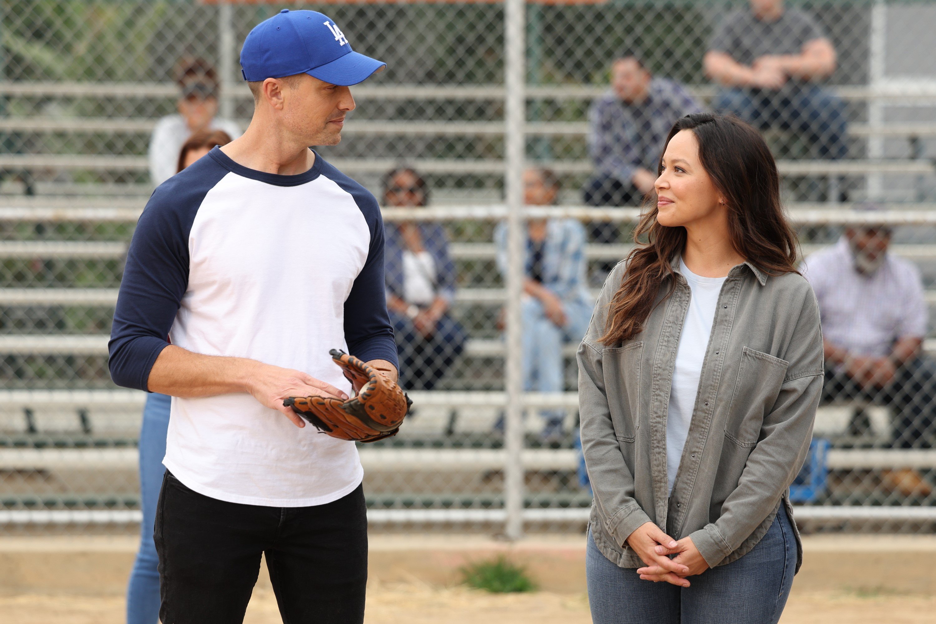 Eric Winter and Melissa O'Neil, in character as Tim Bradford and Lucy Chen in 'The Rookie' Season 5 Episode 11, share a scene on a baseball field. Tim wears a blue and white baseball shirt, black pants, and a blue LA Dodgers baseball hat. Lucy wears a gray button-up shirt over a white shirt and jeans.