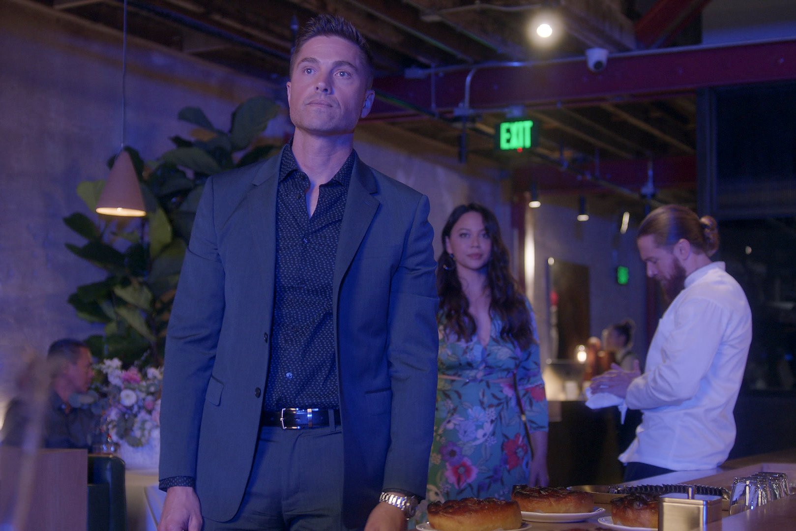 Eric Winter and Melissa O'Neil, in character as Tim Bradford and Lucy Chen in 'The Rookie' Season 5, share a scene at a restaurant. Tim wears a dark gray suit over a black button-up shirt. Lucy wears a light blue dress with pink, purple, and white flowers on it.